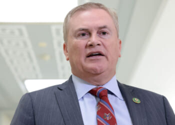 A man in a gray suit, blue shirt, and red patterned tie stands indoors, speaking. He has short light hair and a lapel pin on his jacket. The background is softly lit and out of focus.