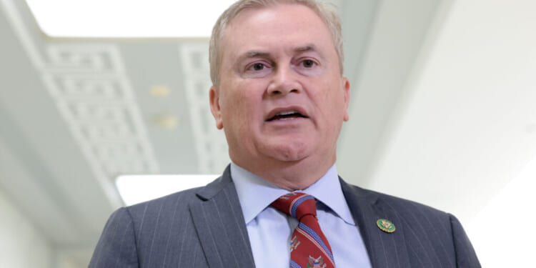 A man in a gray suit, blue shirt, and red patterned tie stands indoors, speaking. He has short light hair and a lapel pin on his jacket. The background is softly lit and out of focus.