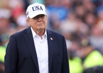 Donald Trump wearing a white USA cap, dark jacket, and white shirt stands outdoors with a blurred crowd in the background.
