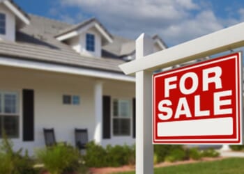 A For Sale sign stands in front of a house planted in the front yard.