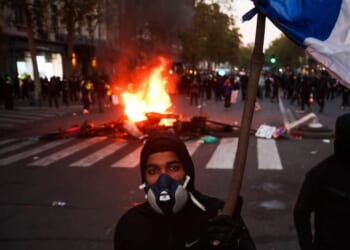 A protestor with a gas mask holds a French flag in front of a fire at the Boulevard des Capucines during a demonstration in Paris on Oct. 17, 2026.