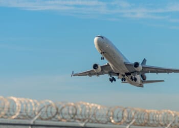 A deportation plane taking off over a barbed wire fence at Miami International Airport.