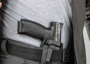 A close-up of a man in a gray t-shirt and jacket revealing a black pistol secured in a tactical belt holster.