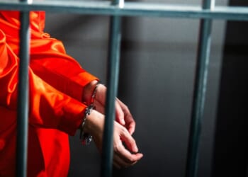 A prisoner wearing an orange uniform sitting in a prison cell.