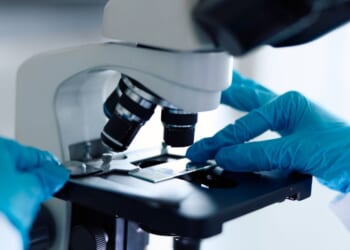 A scientist in a white lab coat and blue gloves carefully placing a sample slide under a microscope.