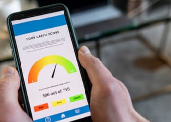 A phone with a credit score displayed on the screen is pictured in a man’s hand.