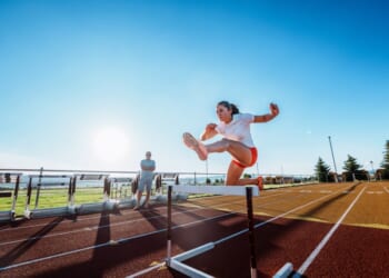 A female collegiate athlete powers over a hurdle at full speed during an intense solo training session with her coach observing nearby.