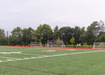 An artificial turf athletic field featuring yard lines, multiple goalposts, a red running track, and a towering stadium light pole.