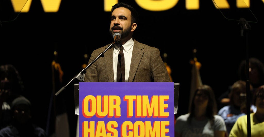 Zohran Mamdani, dressed in a tweed jacket, speaks at a campaign rally.