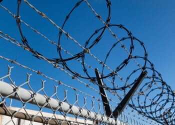 Barbed and razor wire is seen on a fence with a security camera in the background.