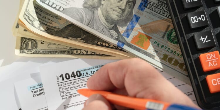 A close-up of the hand of a person filling out a form 1040 on a desk with some dollar bills and a calculator.