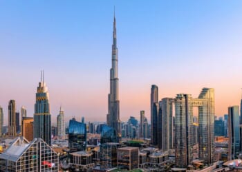 An aerial cityscape of Dubai at sunset with modern skyscrapers, illuminated highways, busy traffic, and dense urban development.