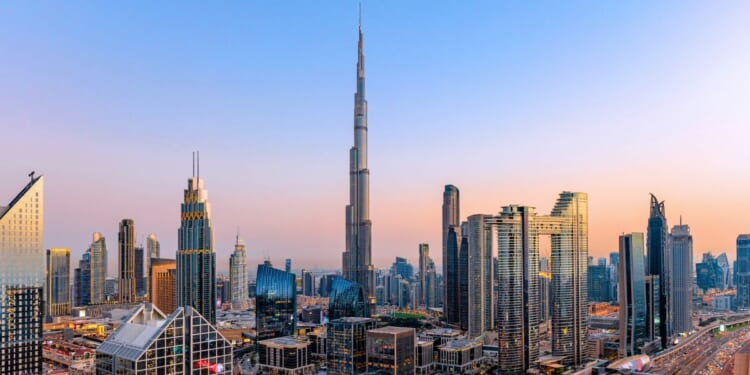 An aerial cityscape of Dubai at sunset with modern skyscrapers, illuminated highways, busy traffic, and dense urban development.