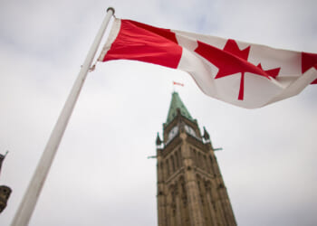 Low angle shot of the Canadian flag with tall, narrow building behind it.
