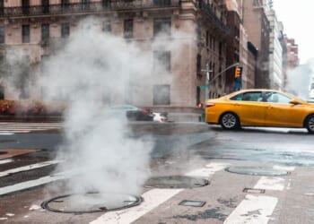 An urban street with steam rising from a manhole and a taxi in motion in New York.