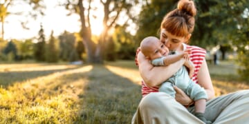 A mother and her baby embrace while sitting down in a meadow in sunset.