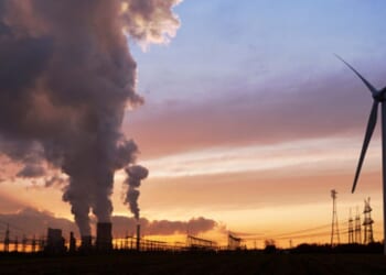 A wind turbine turns as factories pump out smoke in the evening.