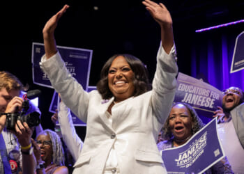 Janeese Lewis George in a white suit waves with both hands in the air as supporters celebrate behind her.