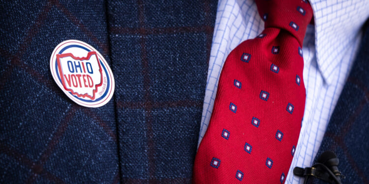 A man's navy suit with red and blue tie features an "OHIO VOTED" sticker.