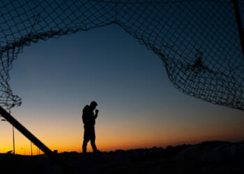 Silhouette of a man at sunrise as seen through a broken border fence.