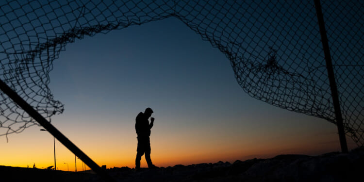 Silhouette of a man at sunrise as seen through a broken border fence.