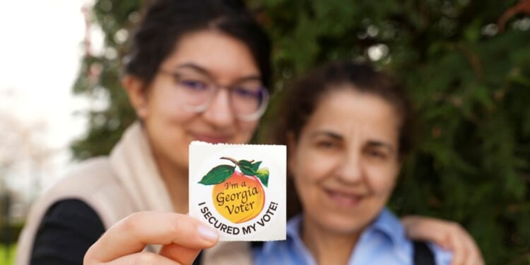 Two women are pictured, while one of them holds a Georgia voter sticker in front of a tree.