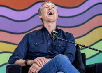 A seated Gavin Newsom with his head back laughing, in front of a backdrop of wavy rows of color.