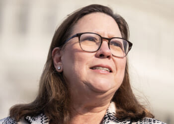 A woman with long brown hair, glasses, and a light-colored jacket stands outdoors in front of a blurred, domed building, looking slightly to the side and smiling.