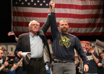 Bernie Sanders lifts Graham Platner's arm at a rally, with American flag as a backdrop.