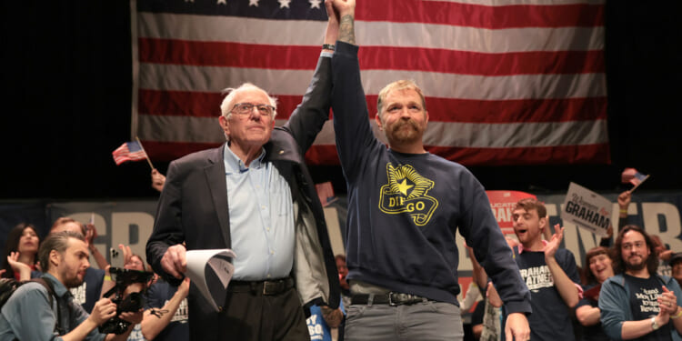 Bernie Sanders lifts Graham Platner's arm at a rally, with American flag as a backdrop.