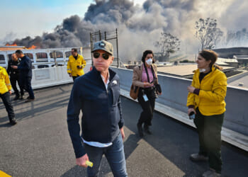 Gavin Newsom, in cap and sunglasses walks as black smoke billows behind him.