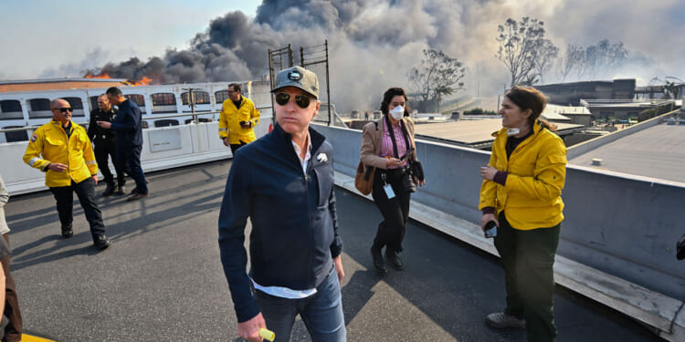 Gavin Newsom, in cap and sunglasses walks as black smoke billows behind him.