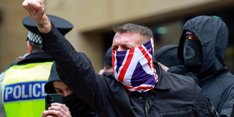 A protester, his face covered in a UK flag, raises his fist as other protesters and a police officer stand behind him.