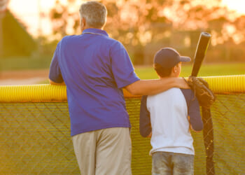 A father in a blue shirt has his arm around his son, who's holding a bat and wearing a baseball jersey and cap. The pair is looking over a ball field in late afternoon.