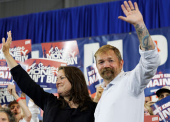 Graham Platner, with his wife by his side, wave to supporters with a campaign banner in the background.