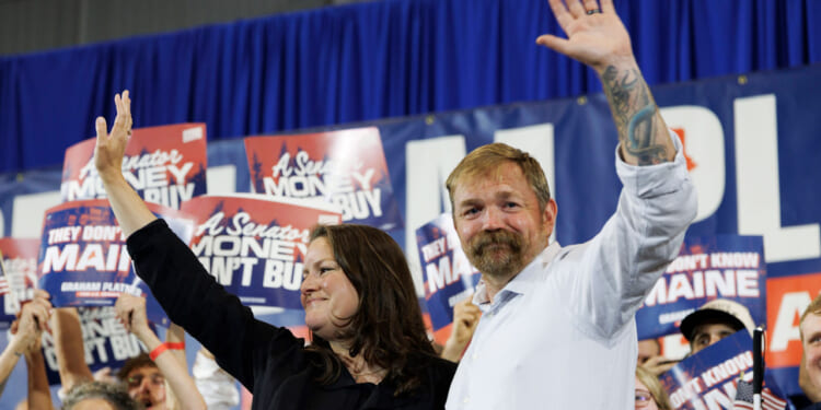 Graham Platner, with his wife by his side, wave to supporters with a campaign banner in the background.