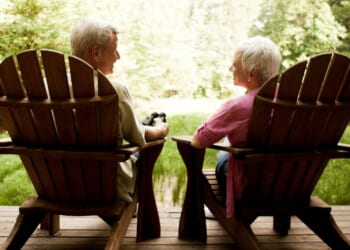 The rear view of a senior couple relaxing in Adirondack chairs on a wooden deck.