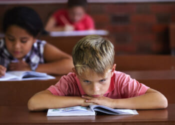 Bored, restless boy rests head on arms as he sits at his classroom desk, while female students behind him are working away.