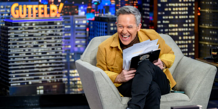 Greg Gutfeld sits in a gray armchair, laughing while holding papers and a clipboard. Behind him is a cityscape backdrop with the word GUTFELD! in bright lights.