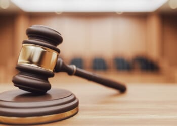 A wooden judge's gavel sits on a desk with a blurred courtroom in the background.