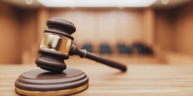A wooden judge's gavel sits on a desk with a blurred courtroom in the background.