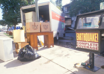 A moving van in a street awaiting furniture to be loaded, a bag of trash and a newspaper vending machine with banner "Earthquake coverage."