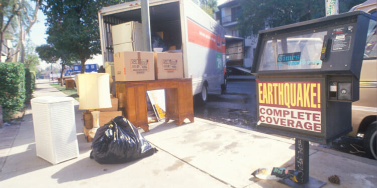 A moving van in a street awaiting furniture to be loaded, a bag of trash and a newspaper vending machine with banner "Earthquake coverage."
