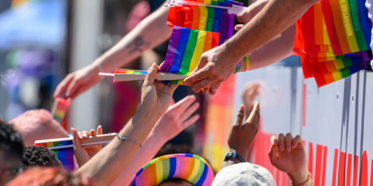 Someone leans over to hand a crowd of people rainbow pride flags during a parade.