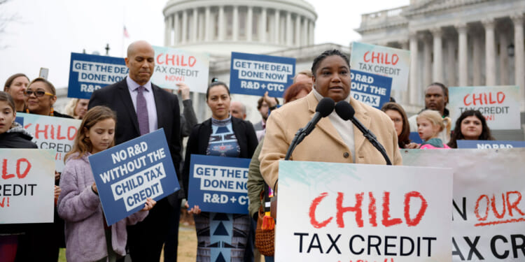 A woman speaks with a "CHILD TAX CREDIT NOW!" sign in front of her and the U.S. Capitol in the background.