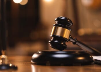 A gavel sits on a wooden block on a wooden desk.