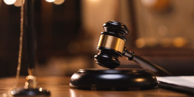 A gavel sits on a wooden block on a wooden desk.