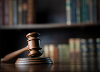 A gavel and wooden block sit on a table while book shelves sit behind them.