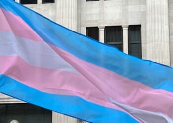 The trans flag waves outside of a building.
