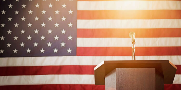 A podium in front of an American flag. Bright light hangs above the microphone.